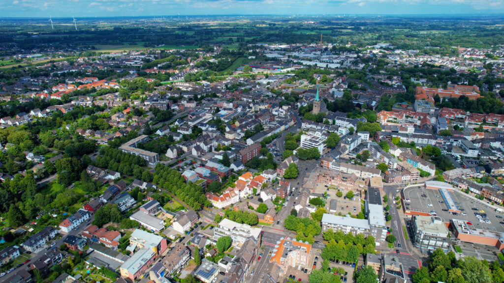 Luftaufnahme einer Stadt mit Häusergruppen, Geschäftsgebäuden, von Bäumen gesäumten Straßen und Grünflächen, umgeben von ländlichen Feldern unter einem blauen Himmel mit vereinzelten Wolken.