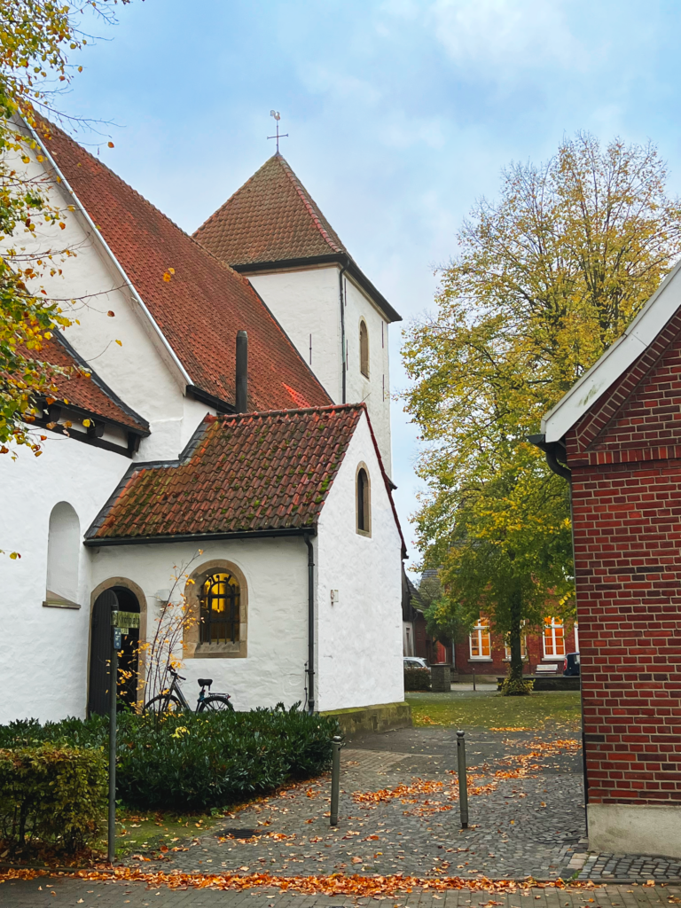 An einem Herbsttag steht eine weiße Kirche mit rotem Ziegeldach neben einem kleinen Backsteingebäude. Laub liegt verstreut auf dem Boden, im Hintergrund sind Bäume mit gelbem Laub zu sehen.
