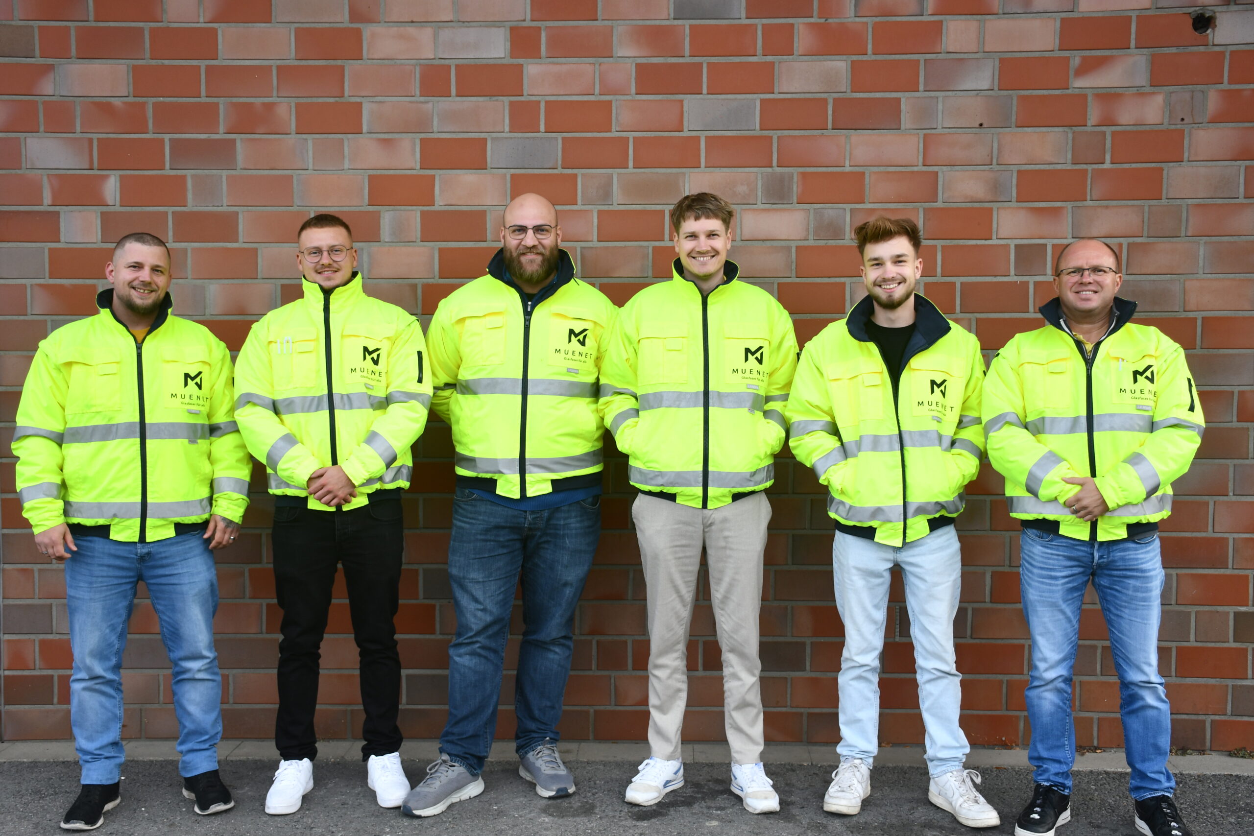 Sechs Männer stehen Seite an Seite vor einer Backsteinmauer, alle tragen leuchtend gelbe Warnwesten mit demselben Logo und lächeln in die Kamera.