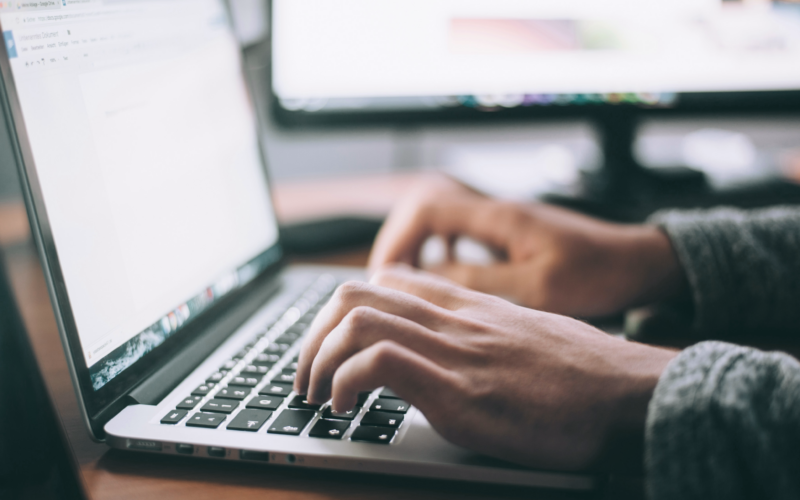 Nahaufnahme einer Person, die an einem Schreibtisch auf einer Laptop-Tastatur tippt. Im Hintergrund ist ein Computermonitor zu sehen. Der Fokus liegt auf den Händen und dem Laptop, was darauf hindeutet, dass die Person gerade arbeitet oder studiert.