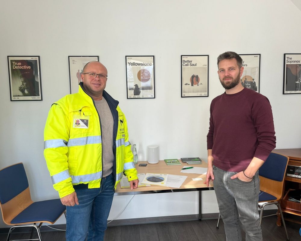 Viktor Kornewald aus dem MUENET-Team und Tim Ehrlich von der Breitbandkoordination der Verbandsgemeinde Forst stehen in einem Raum mit vier gerahmten Serienpostern an der Wand. Kornewald trägt eine gelbe Warnjacke, Ehrlich ein rotes Langarmshirt. Auf dem Tisch liegen Papiere und Broschüren.